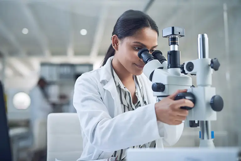 A woman wearing a lab coat focuses on her work, peering through a microscope in a scientific laboratory environment.