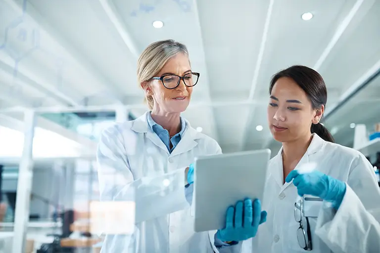 A group of individuals in lab coats are engaged in discussion while holding tablets in a laboratory setting.
