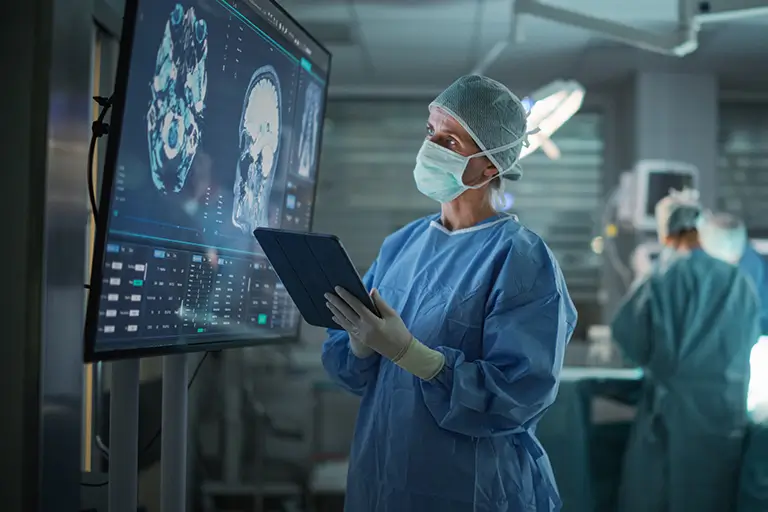 A surgeon in scrubs examines a tablet while standing in a hospital setting, focused on patient information.