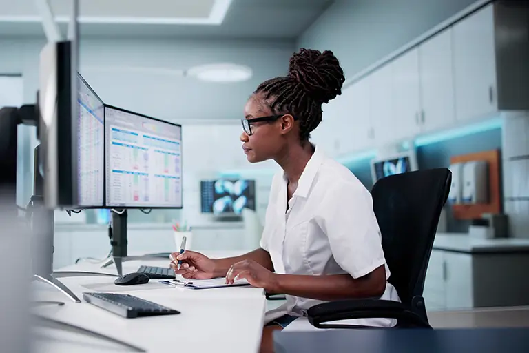 A woman in a white lab coat focused on her work at a computer in a laboratory setting.